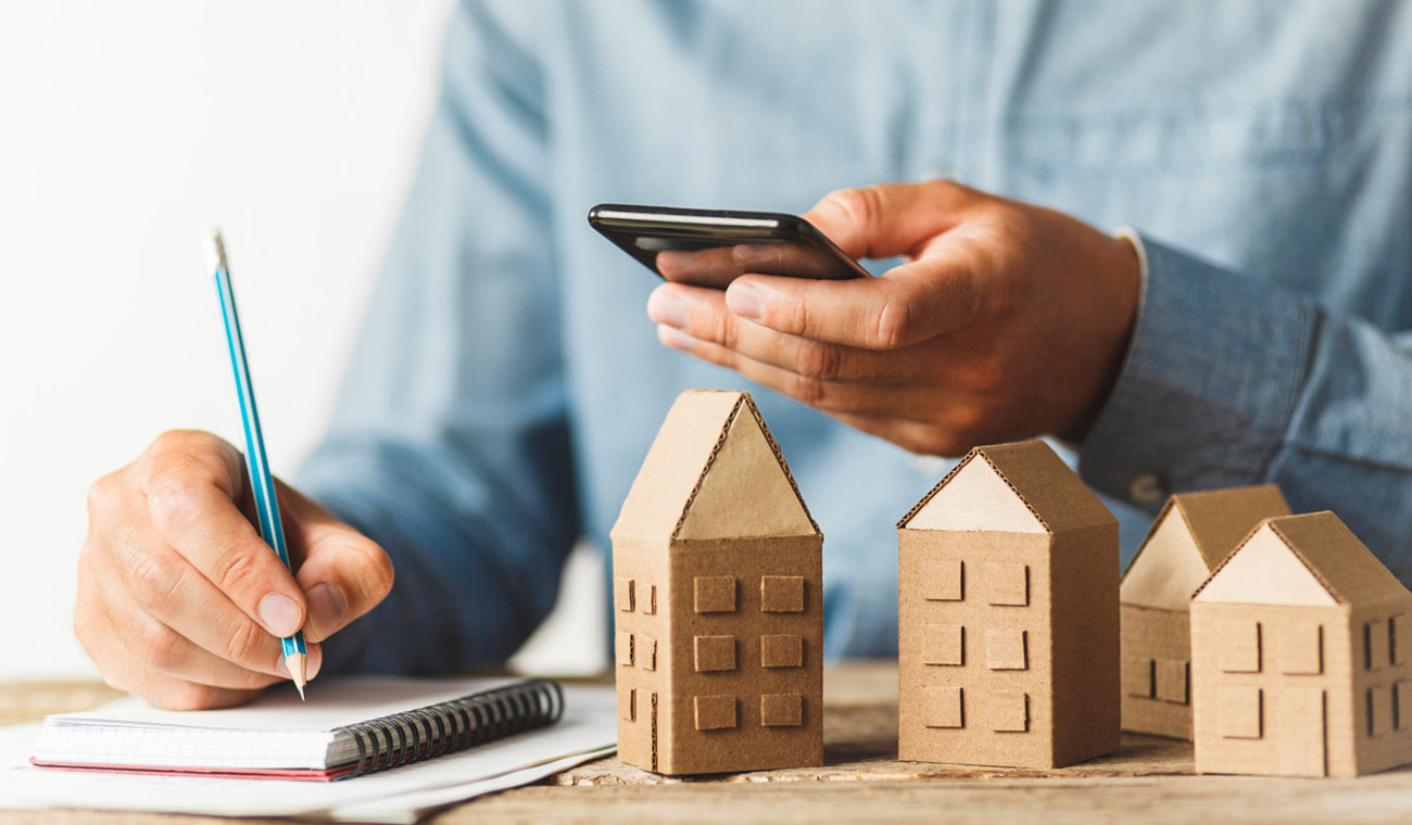 man using his phone to make calculations while writing on a notebook next to miniature cardboard homes